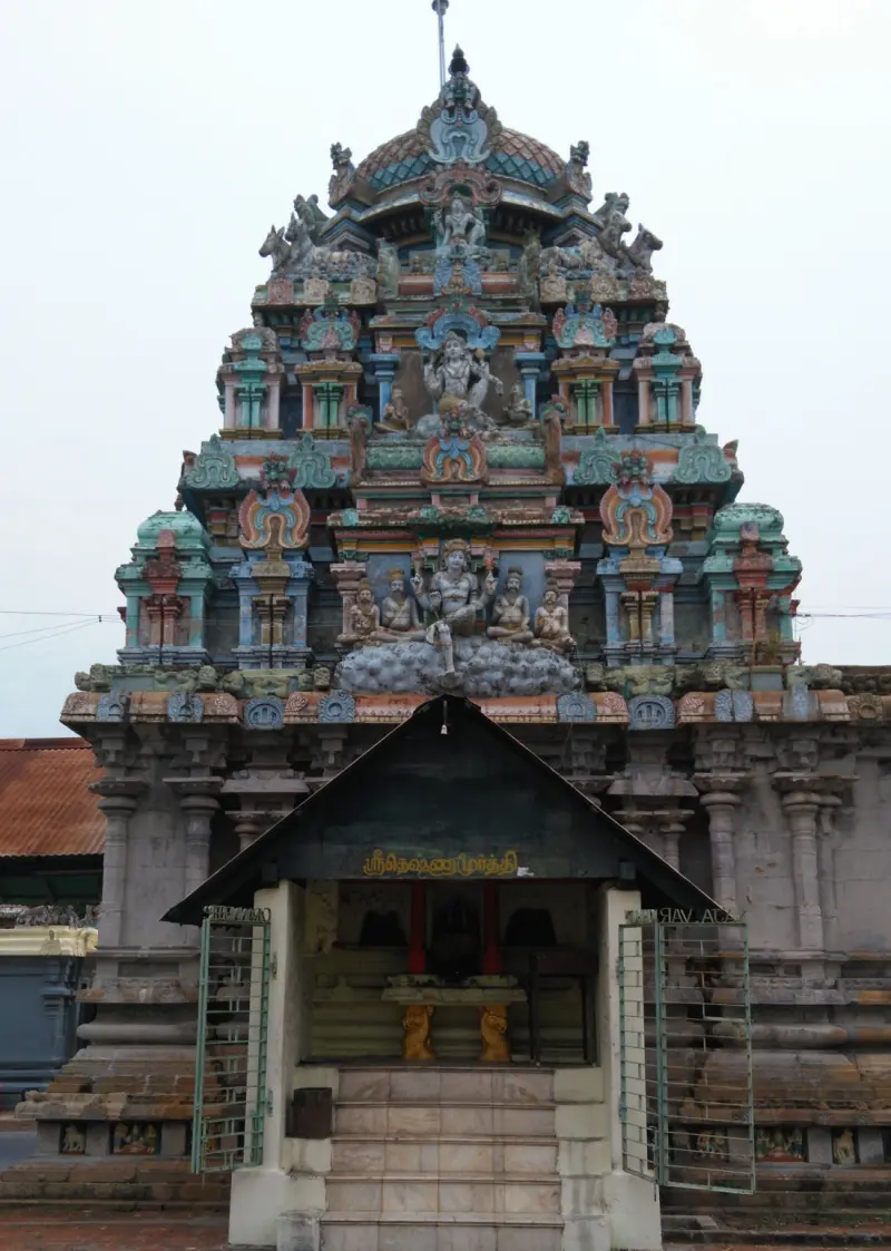 Small open shrine with a Tamil name board and steps, set against a carved vimanam with many figures.