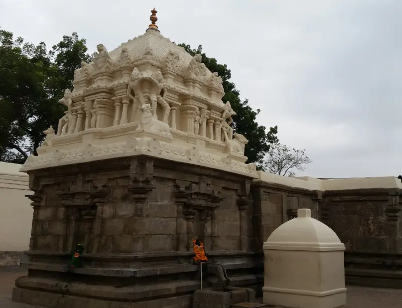 A stone shrine with a cream-coloured tower of carved figures, a small domed structure beside it, and trees behind.