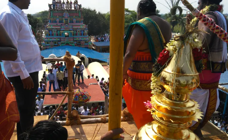 Priests stand on a high platform beside a gilded kalasam, with a crowd and a painted tower beyond.