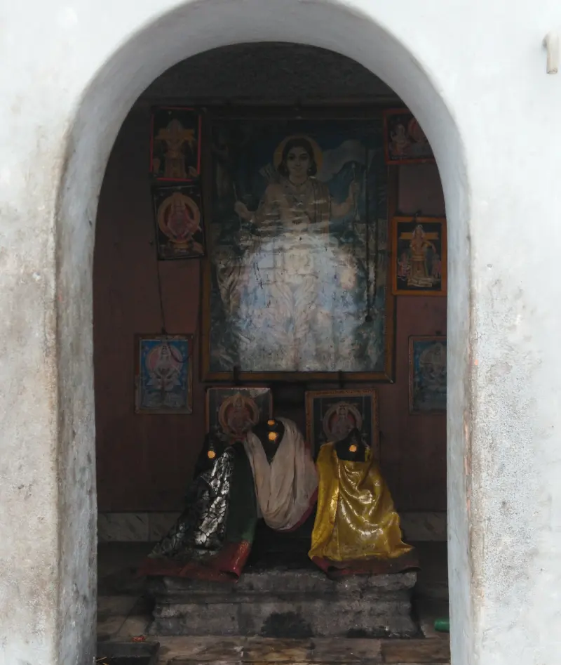 Through a white arch, three idols in cloth on a stone base, with framed pictures on the wall behind.