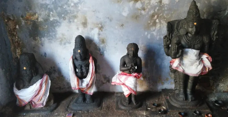 Four black stone idols in white cloth stand along a wall, with small lamps at their feet