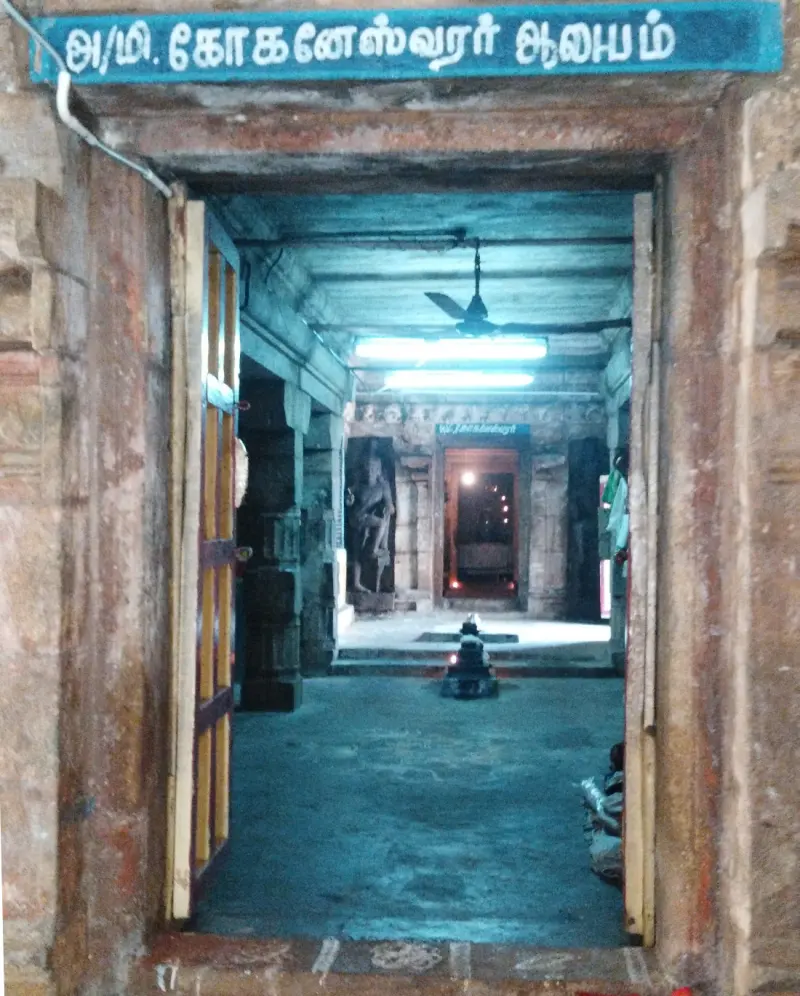 View through a stone doorway under a blue Tamil name board, down a hall to a lit inner shrine