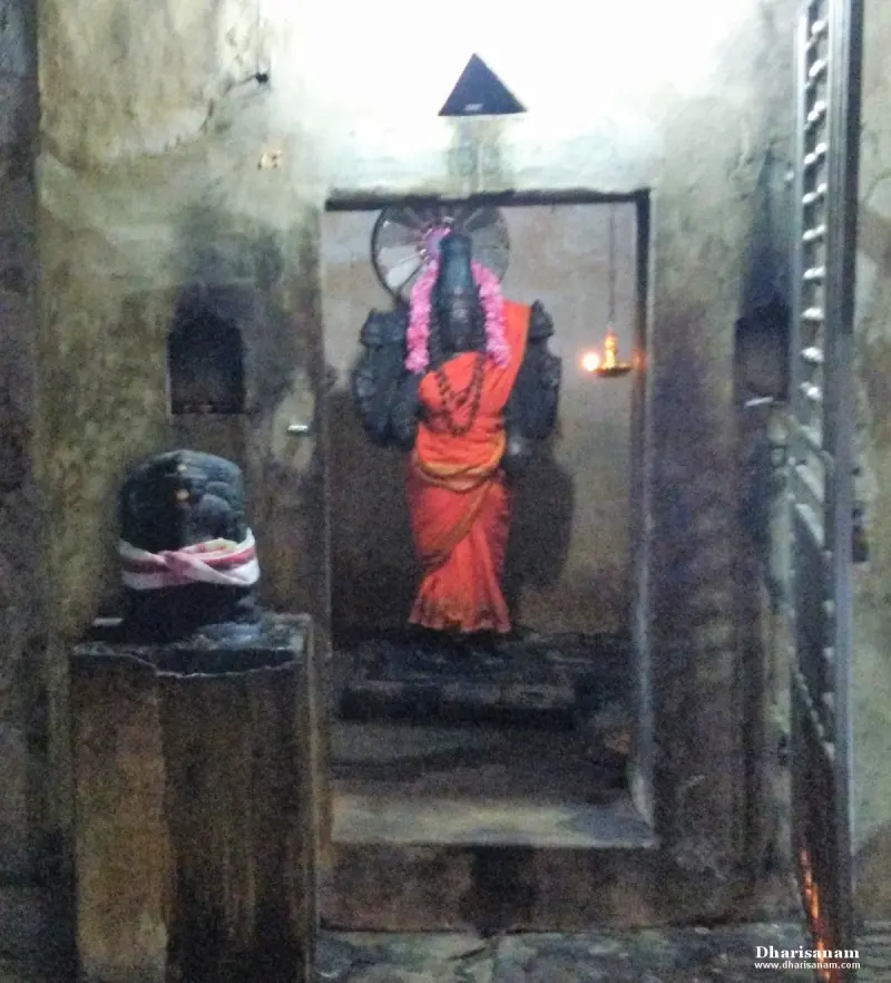 A dark stone idol in an orange cloth and flower garland stands in a niche, with a Shivalingam on the left