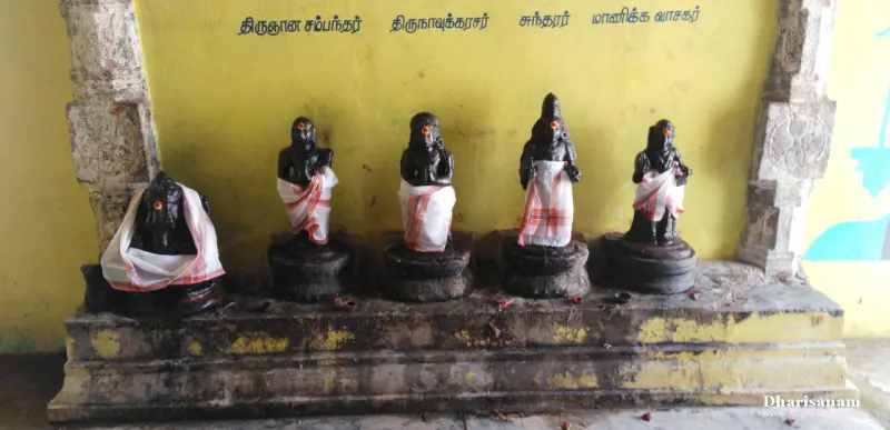Five small dark stone figures in white and orange cloth in a row on a stone platform, with names on the wall