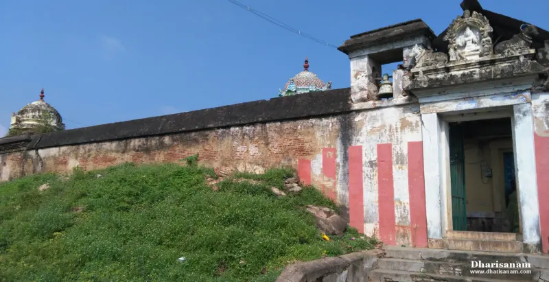 A long brick wall beside a grassy slope, with a gateway topped by a Ganesha figure at right and two domes beyond.