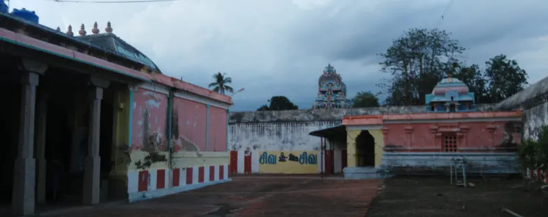 A paved courtyard with painted pink and yellow walls lettered in Tamil, small shrines and a tower behind.