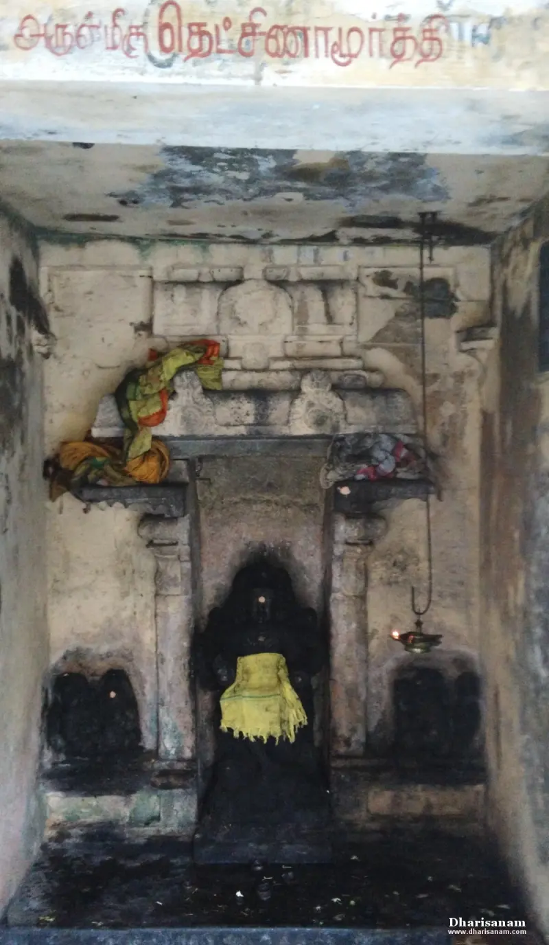 A dark stone figure draped in a yellow cloth in a small niche, with a hanging lamp and Tamil lettering above