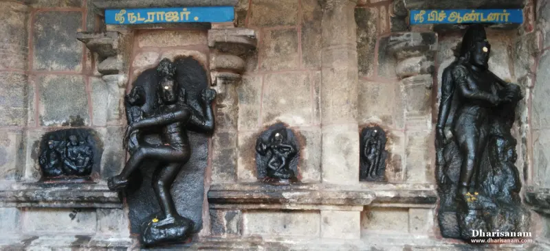Two dark stone figures on a wall with blue Tamil name boards, with small carvings between them
