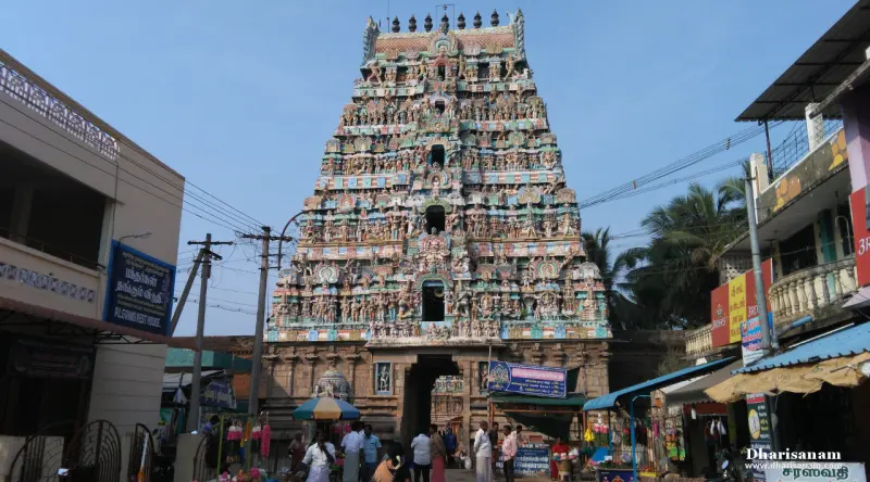 A tall tower covered in colourful sculptures stands above shops and houses on a narrow street