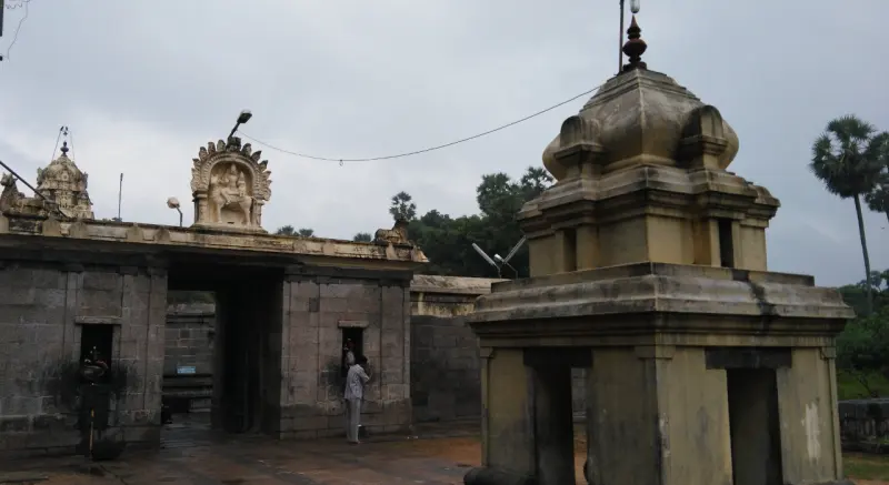 A stone gateway with a carved sculpture of two seated figures on top, and a small domed shrine at the right.
