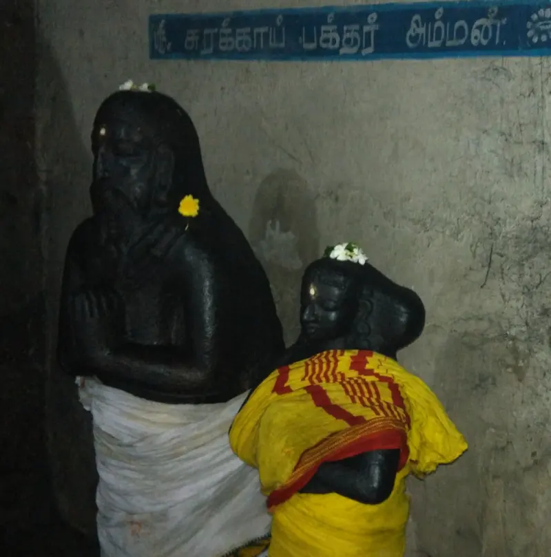 Two dark stone idols, a bearded man with folded hands and a woman in a yellow cloth, with garlands of flowers.