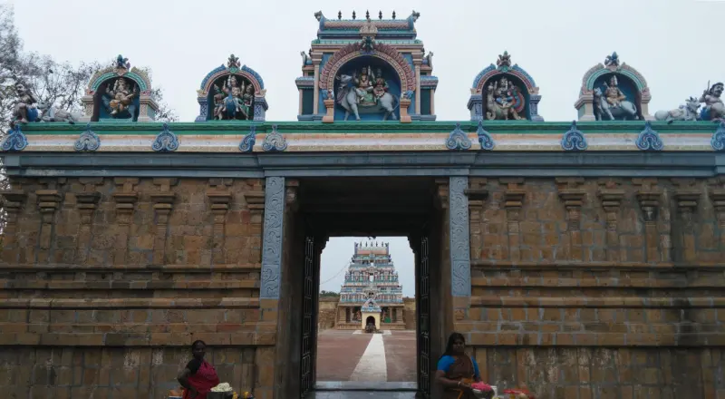Painted figures above a stone gateway; two visitors stand at its foot and a tower shows ahead