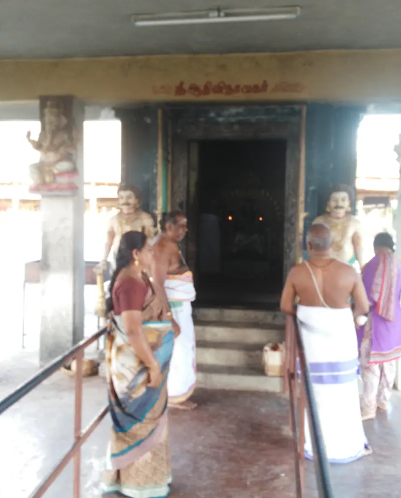 Devotees stand before a dark shrine doorway with a Tamil sign for Aadhi Vinayakar; guardian figures flank the steps.