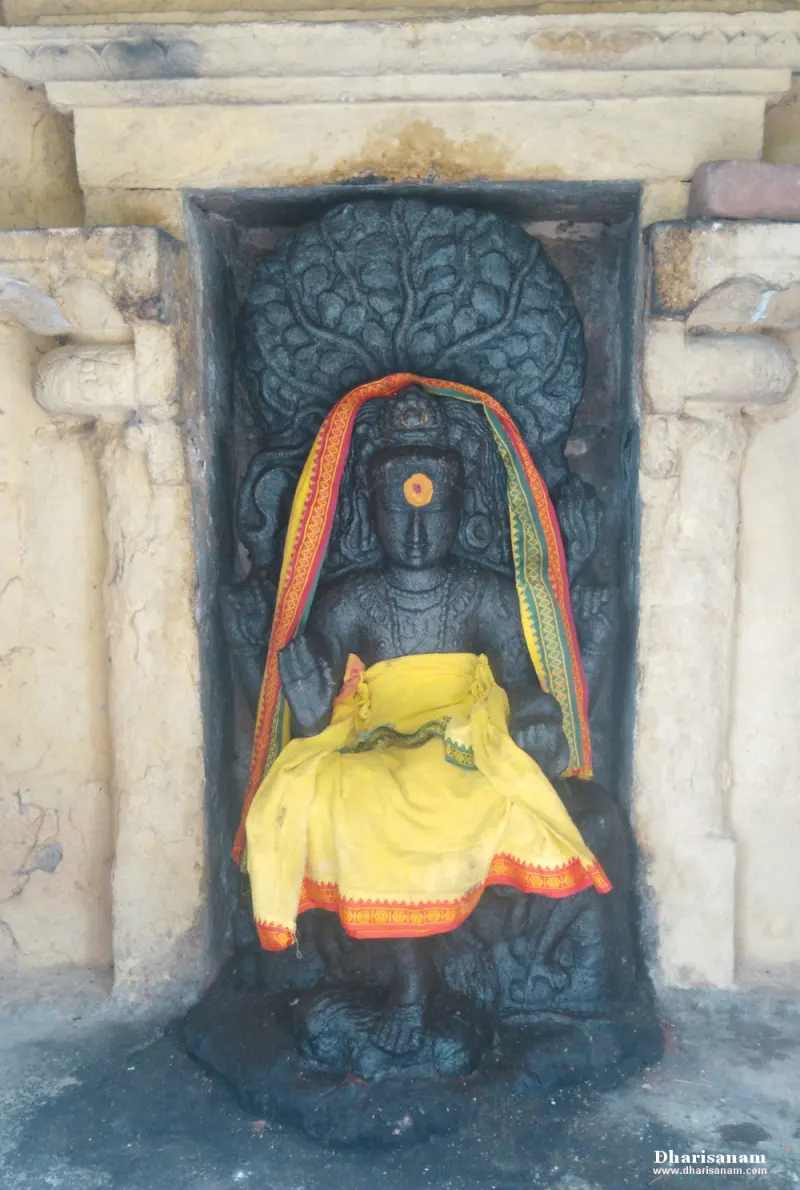A seated dark idol in yellow cloth and a shawl, set in a niche with a tree carved behind.