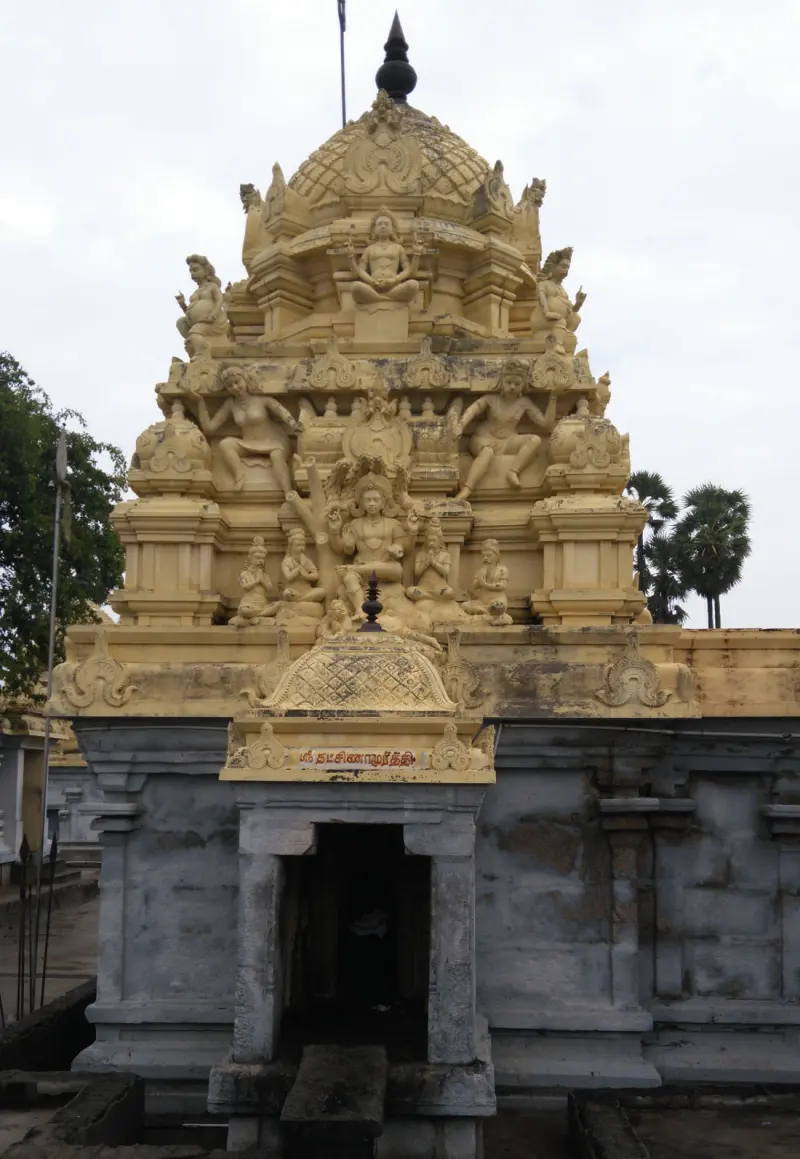 A shrine with a tall carved tower full of figures, and a name board for Dakshinamurthy over the open doorway.
