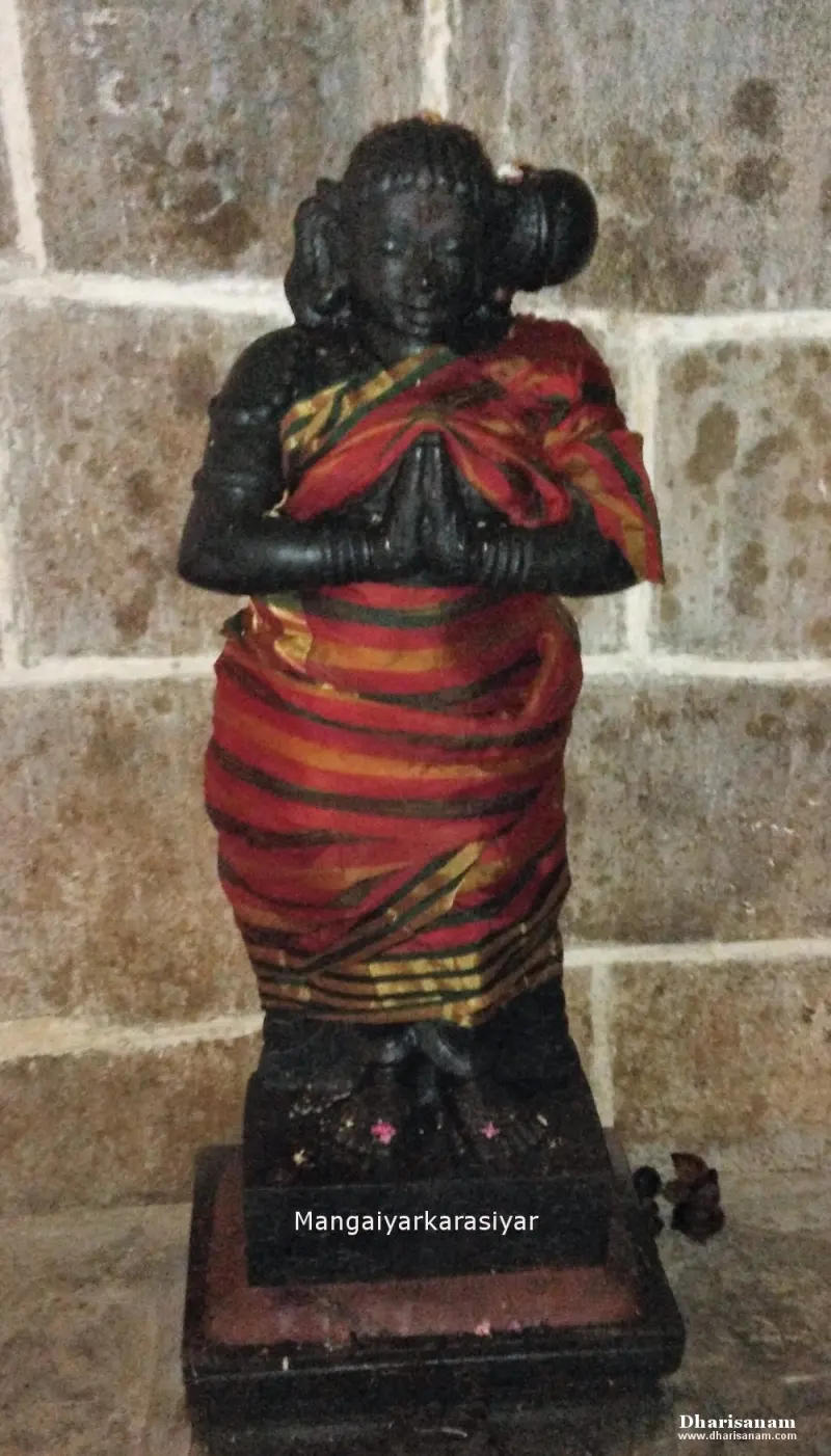 Black stone figure with folded hands in a red sari, standing before a stone wall, with a name label on the base