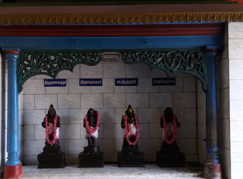 Four dark figures with red flower garlands under blue Tamil name boards, beneath a carved wooden arch