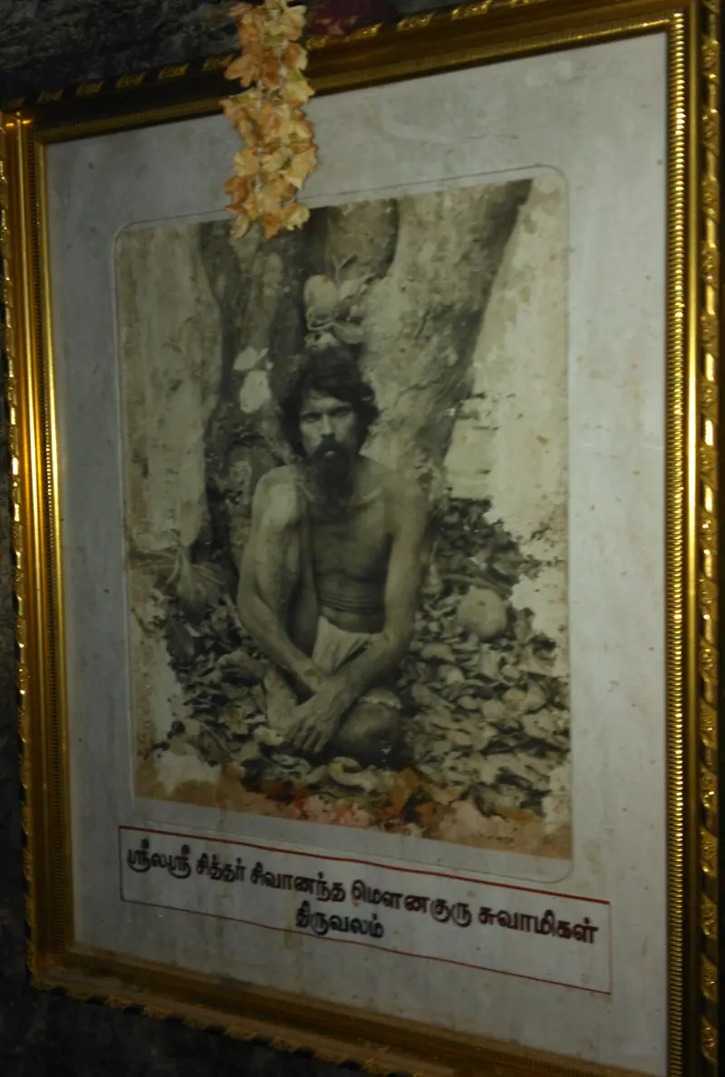 An old framed photograph of a bearded man seated on leaves, with a Tamil name label below