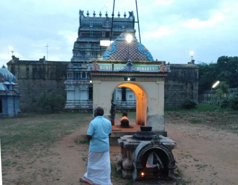 A man in a blue shirt and white dhoti stands before a small lit shrine, with the tower and temple wall beyond.