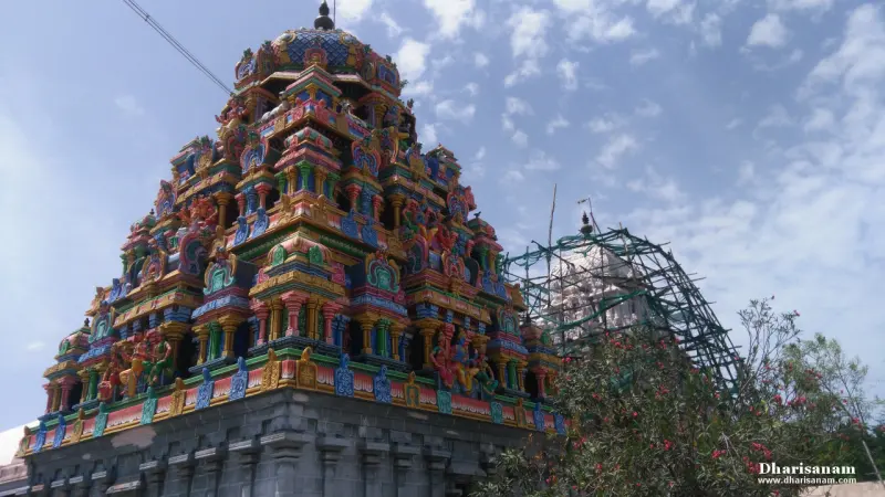 A tiered painted tower rising above a grey stone base, with scaffolding at right and a flowering bush