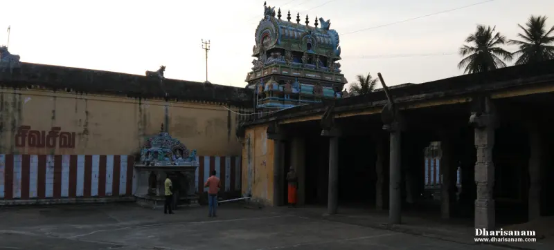 A small carved shrine by a striped wall, with a tower behind it and a pillared hall at right