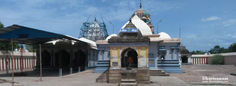 A small stone shrine with a name board in the foreground, a blue-roofed open hall at left and white domes behind