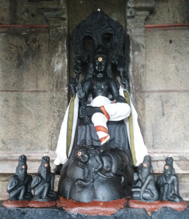 A black stone Dakshinamurthy seated under an arch, with four small seated sages at his feet.