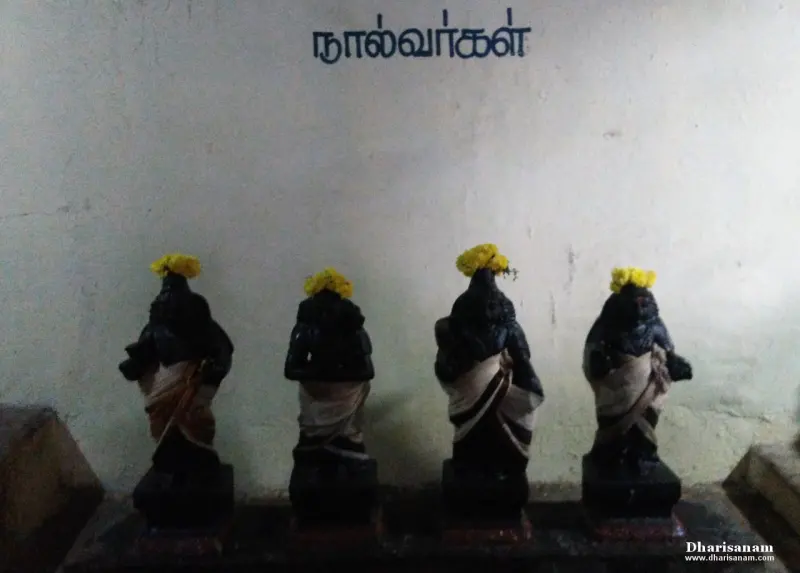 Four small dark idols with yellow flowers on their heads, standing before a wall that says Nalvargal in Tamil