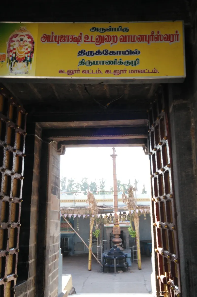 A doorway with open wooden gates under a yellow Tamil name board, and a flagstaff in the courtyard beyond