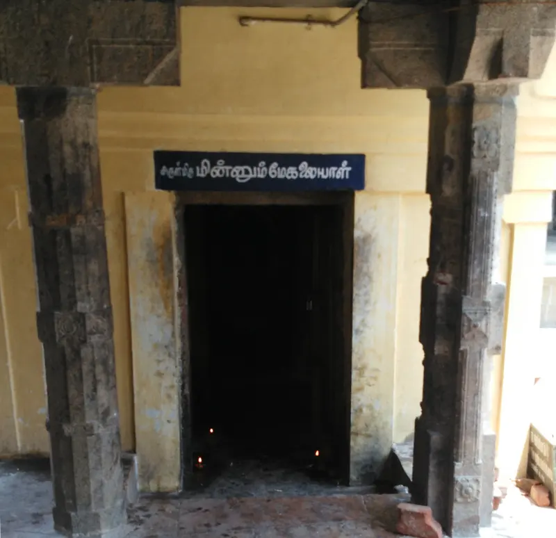 A dark doorway with a blue Tamil name board and small lamps, between stone pillars and a yellow wall