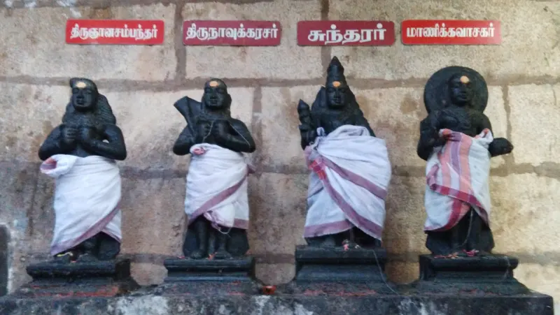 Four black stone saints in white cloth stand in a row on a pedestal, each under a red Tamil name plate.
