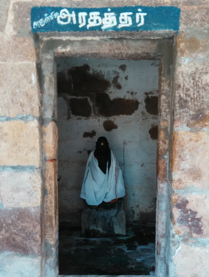 A small stone doorway with a Tamil name board above it; inside, a dark idol draped in white cloth.