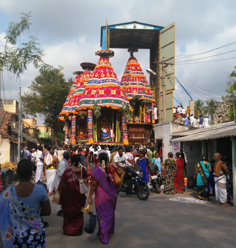 A tall temple car with colourful cloth canopies, surrounded by a large crowd in a narrow street