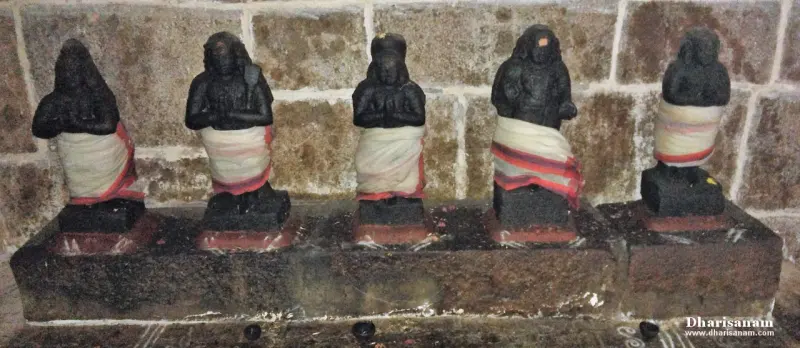 Five small black stone idols wearing white and red cloth, seated in a row against a stone wall