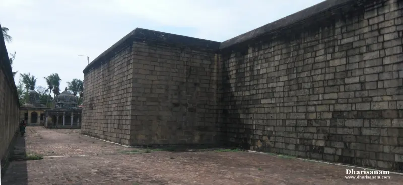 A tall stone block wall running along a paved path, with small pillared structures far at the left