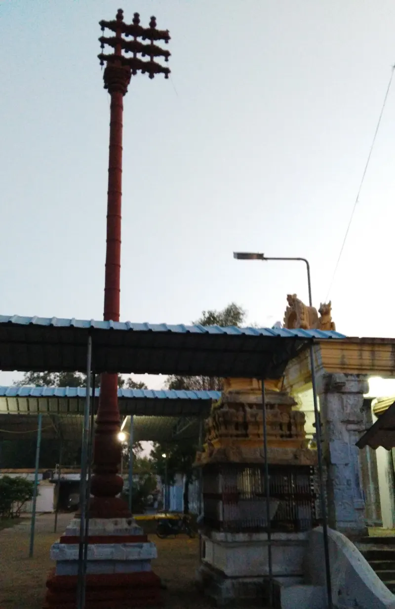 A tall red flag post on a stepped base beside a covered walkway and a small stone shrine.