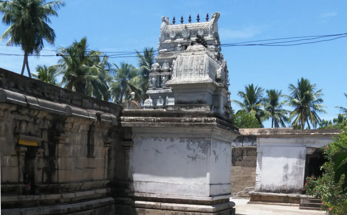 A small stone tower with carved figures rising above a whitewashed wall, with coconut palms behind it.