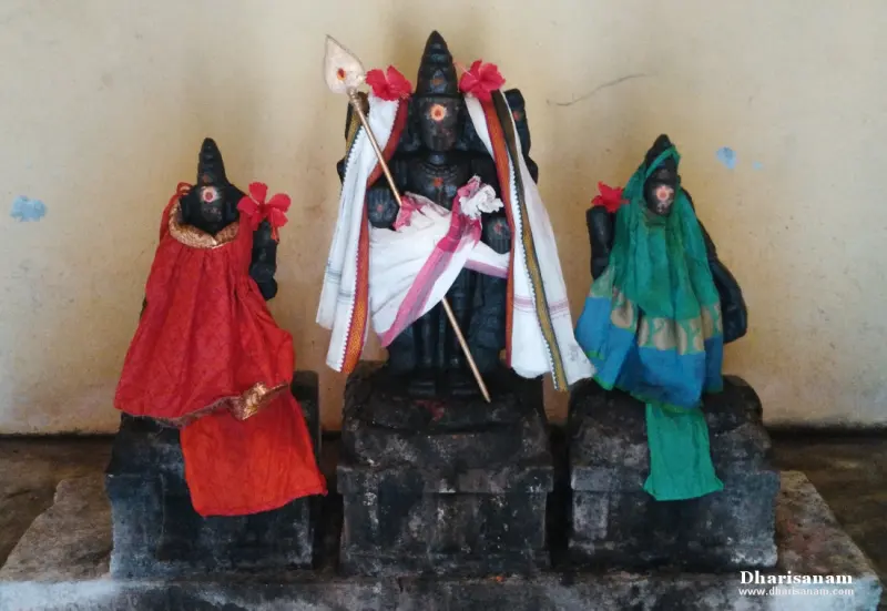 A dark stone Murugan holding a spear, with two consort idols in red and green silk on either side.