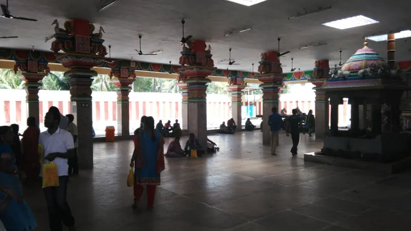Colourful carved pillars line a wide stone hall where devotees walk and sit; a small domed shrine stands at right.