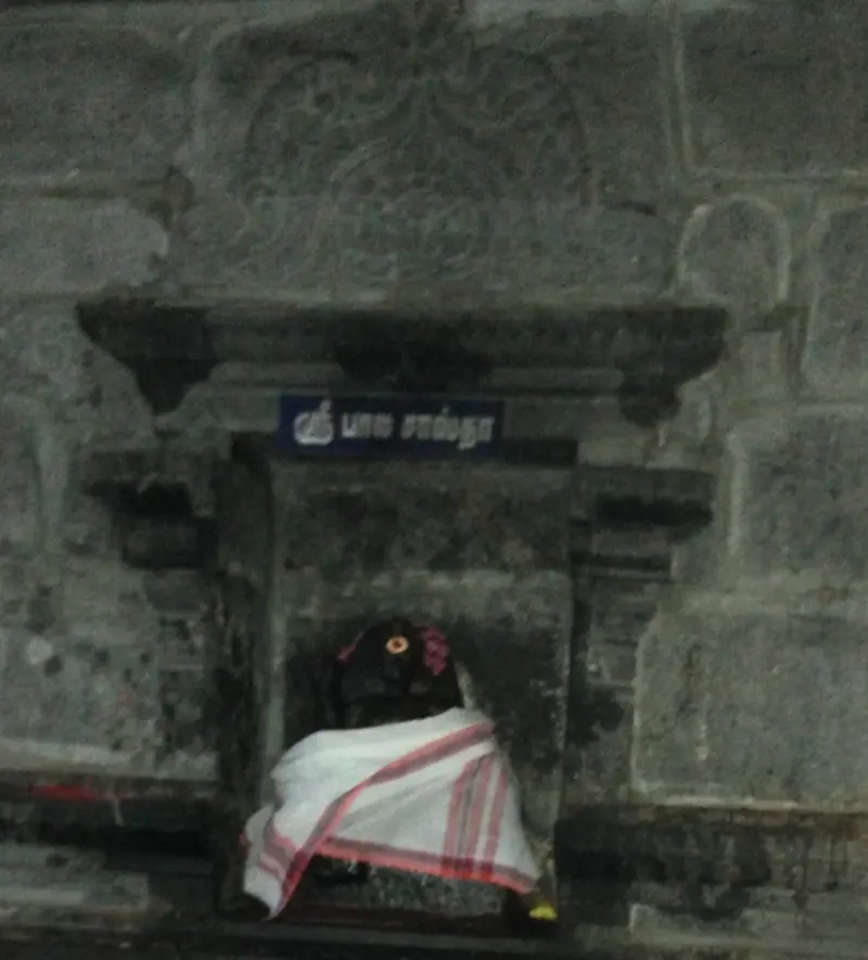 A small niche in a stone wall with a blue name board and a dark idol draped in a white cloth with a red border