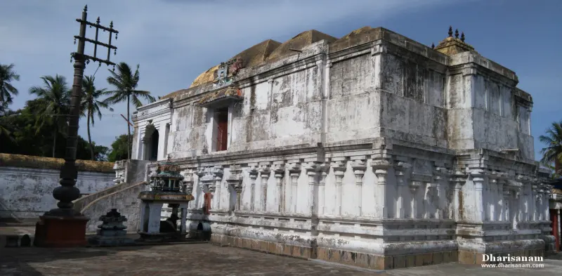A large whitewashed shrine with pillared walls stands beside a tall flagstaff under a blue sky with palm trees