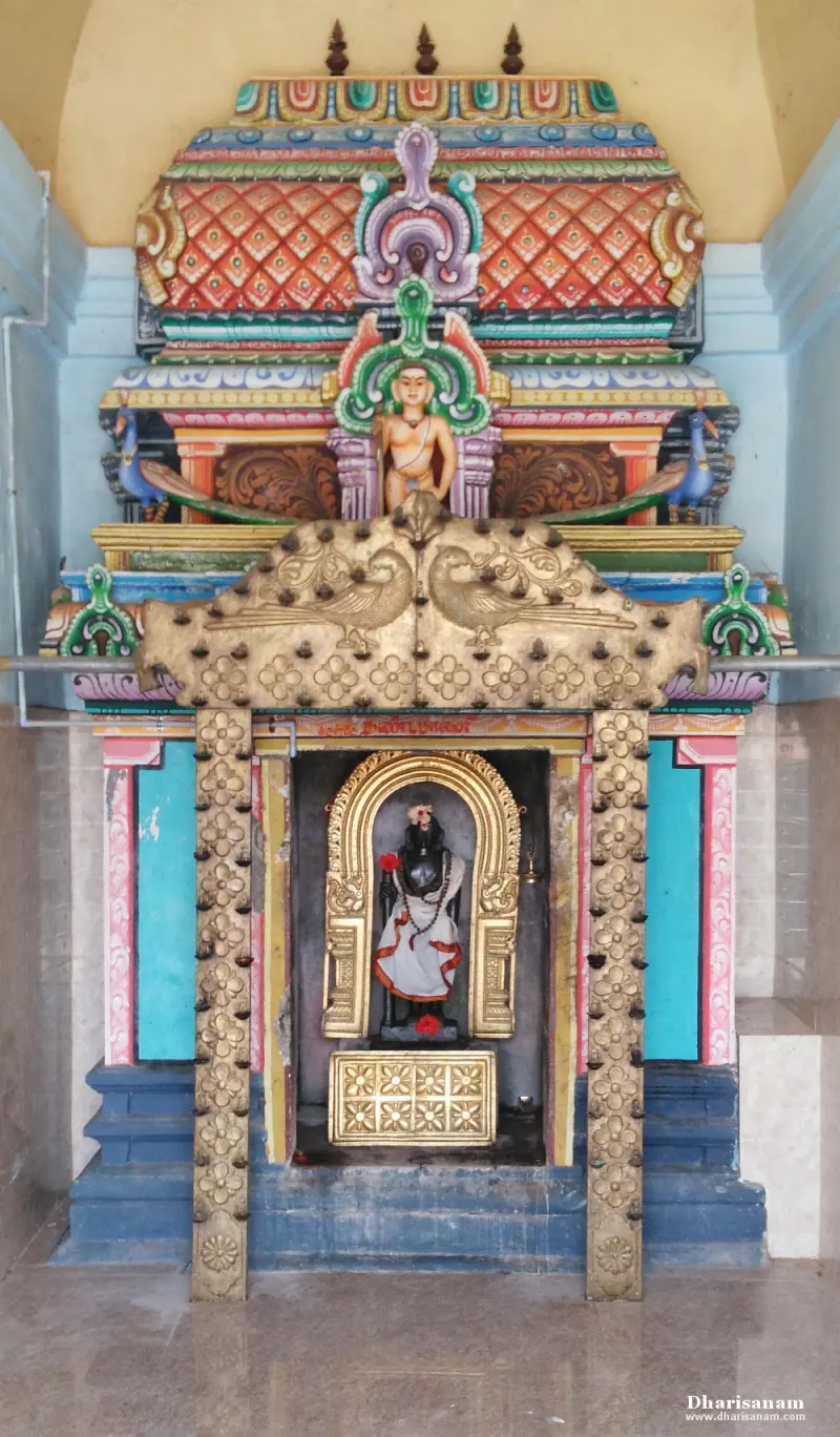 A shrine with a gold-coloured arch and peacocks above, and a standing idol with a red flower inside.