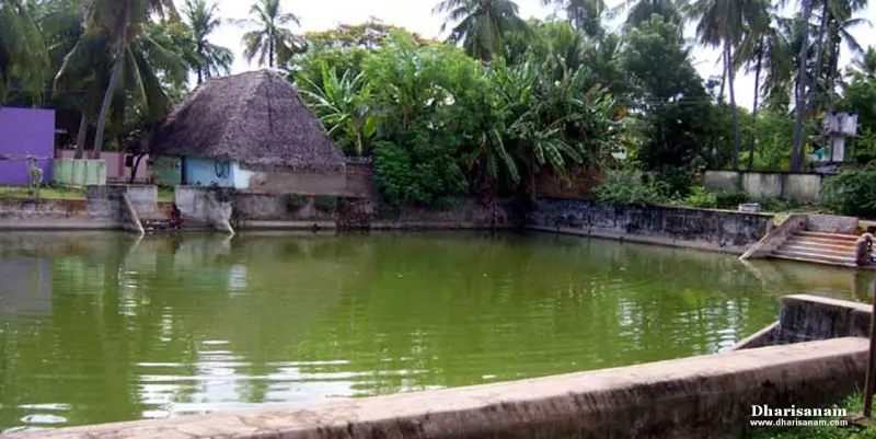 A green-watered tank with stone steps at the right and a thatched hut beyond the far bank
