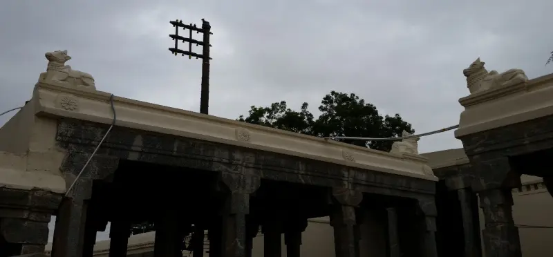 A dark flagstaff rising behind a stone hall roof with Nandi figures on its corners, against a grey sky.