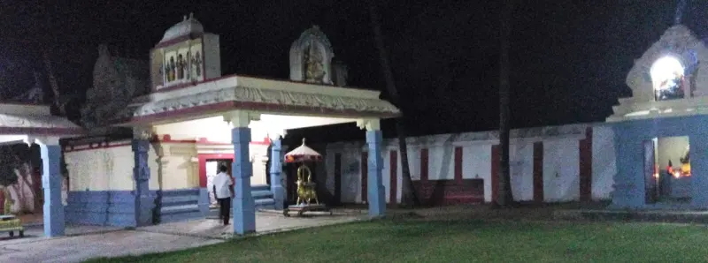 A pillared porch with a small gold-coloured figure inside, beside a white compound wall and gateway