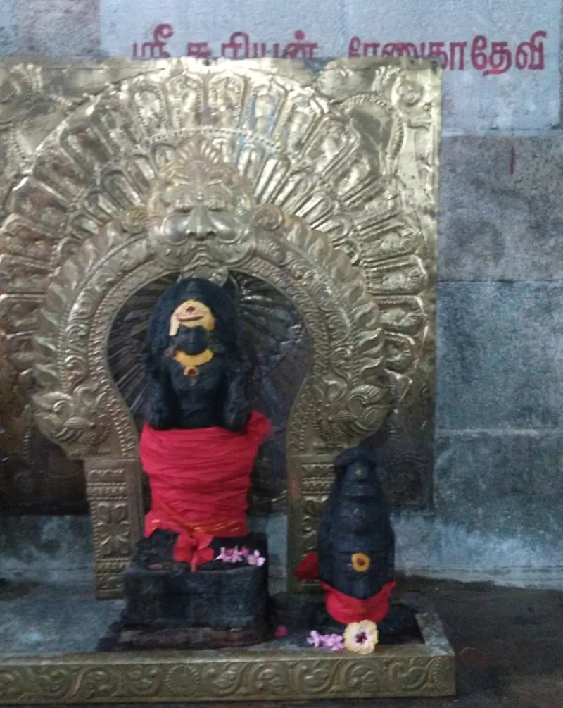 A dark idol in red cloth under a gold arch, with a small second idol beside it and a name board above.