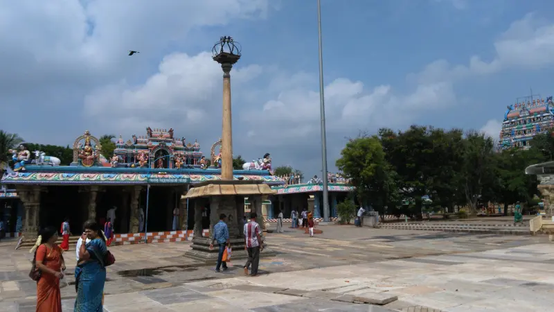 A tall stone pillar in a wide paved courtyard, with a painted mandapam and visitors walking nearby.