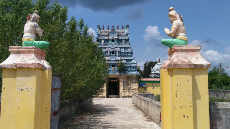 A paved path between two yellow pillars topped by seated figures, leading to the gopuram ahead