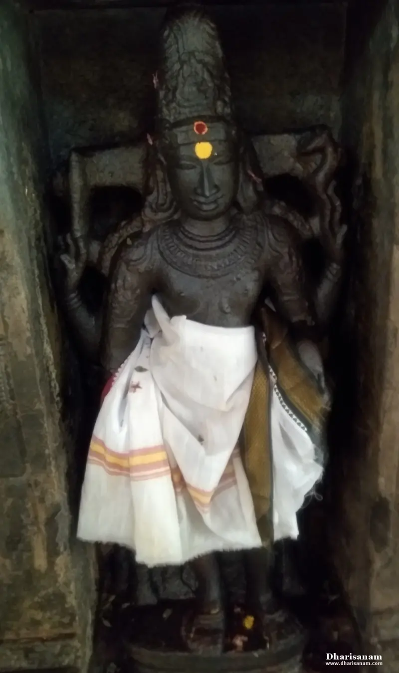 A tall dark stone idol with four arms and a high crown, wearing a white cloth, in a wall niche