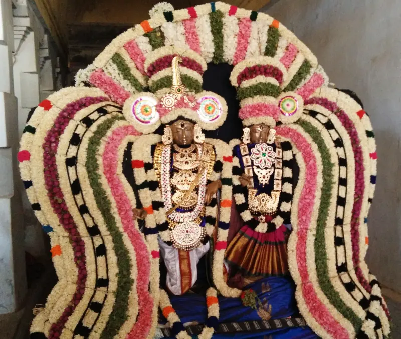 Two festival idols with gold jewellery, framed by an arch of flower garlands in pink, white and green.